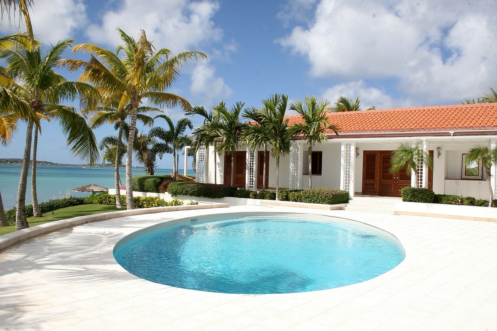 A Jumby Bay residence — terracotta-tile roof, beachfront elevation with timber doors and rounded forecourt pool. Built by Elderfield Gibbs Smith, Master Builder.